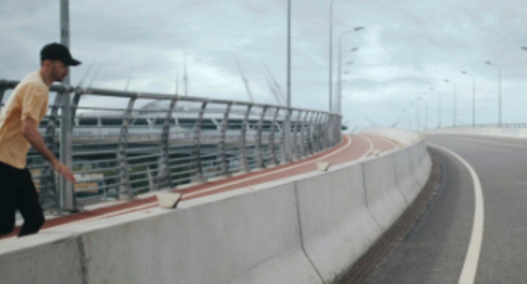 A man in a yellow shirt and cap walks along a curved, empty overpass on a cloudy day.
