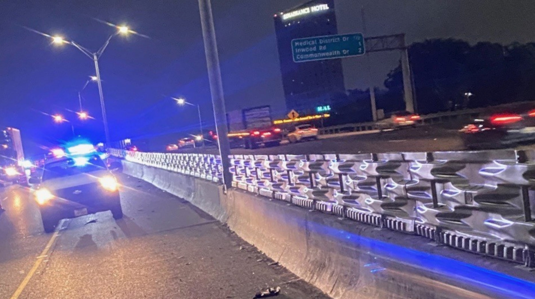 Police car with flashing lights on a highway at night; traffic and road signs visible in the background.