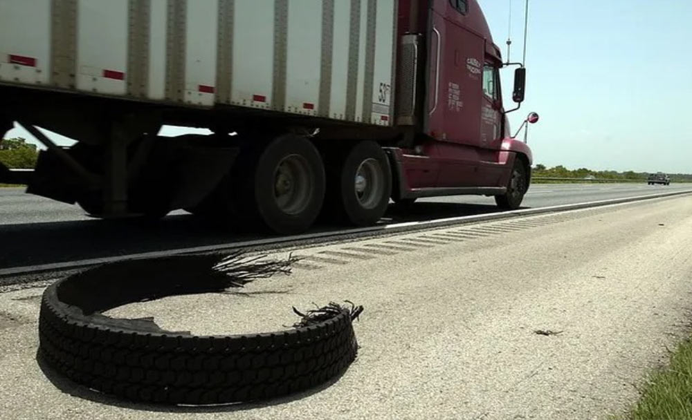 A shredded truck tire lies on the shoulder as a semi-truck drives past on the highway.