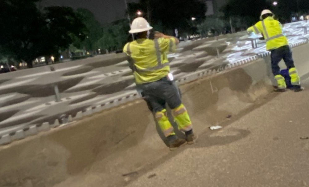 Two construction workers in safety gear stand by a barricade at night, working on a road project.