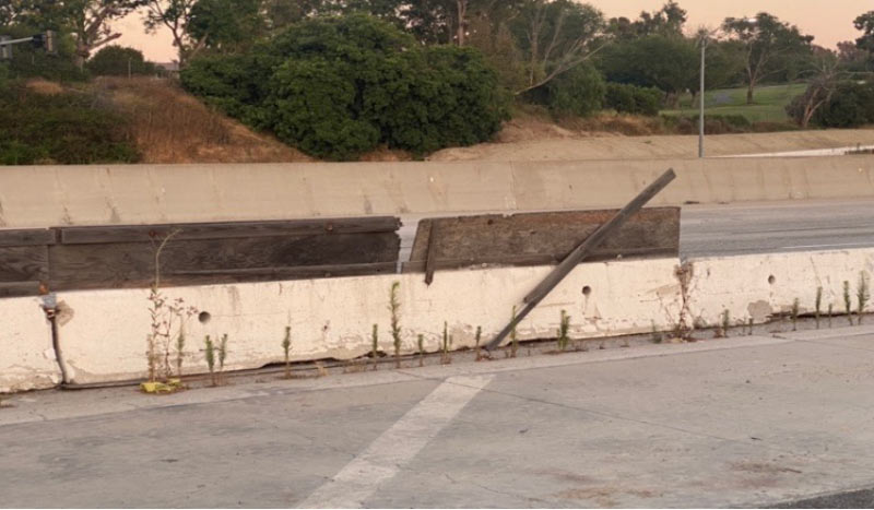 Concrete highway barriers with wood planks and weeds growing at the base, beside an empty road.