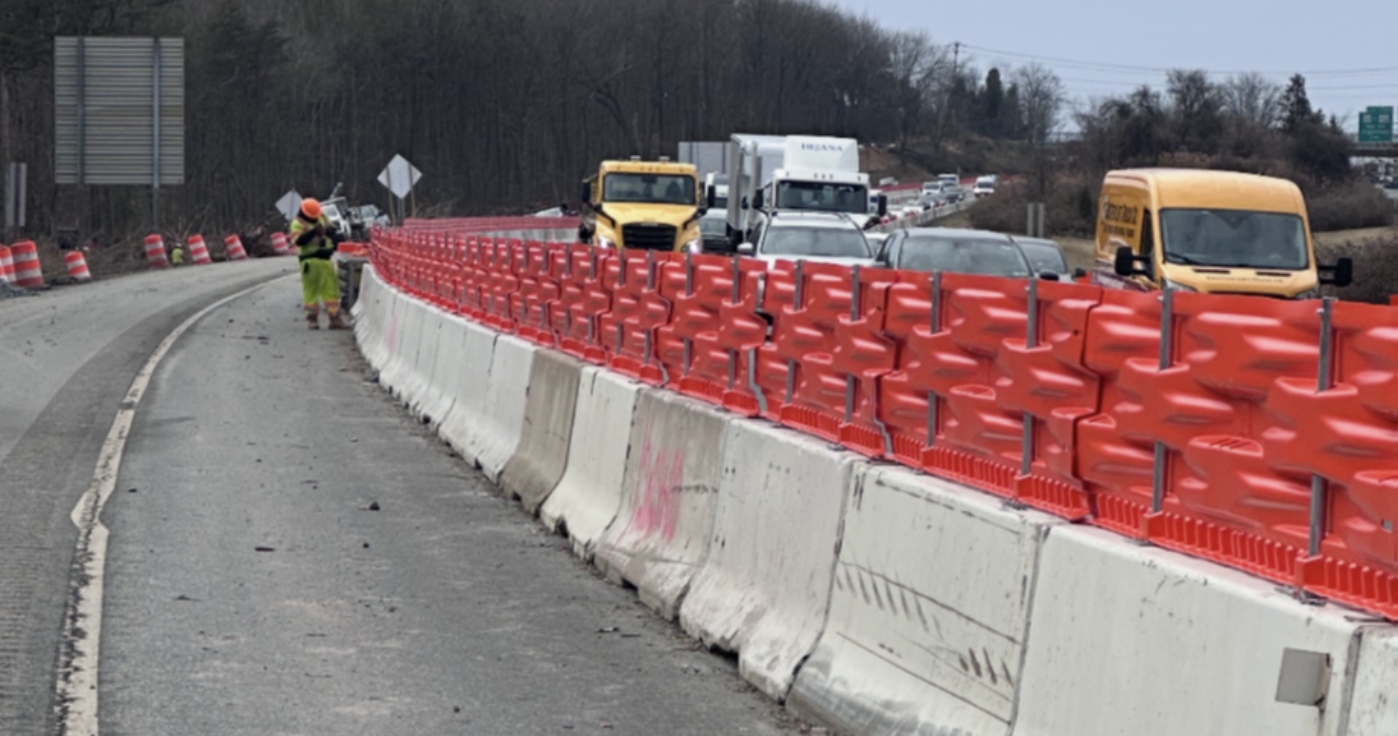 Construction workers near traffic barriers on a highway with cars and trucks in a traffic jam behind them.