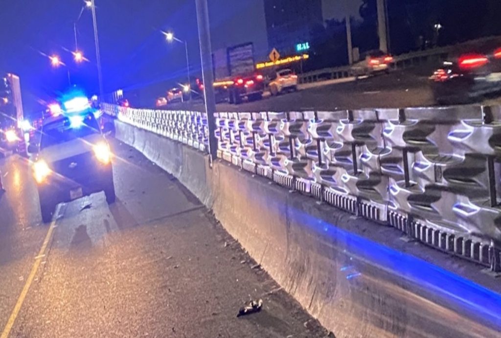 Police car with flashing lights blocks a highway lane at night; traffic continues in other lanes.