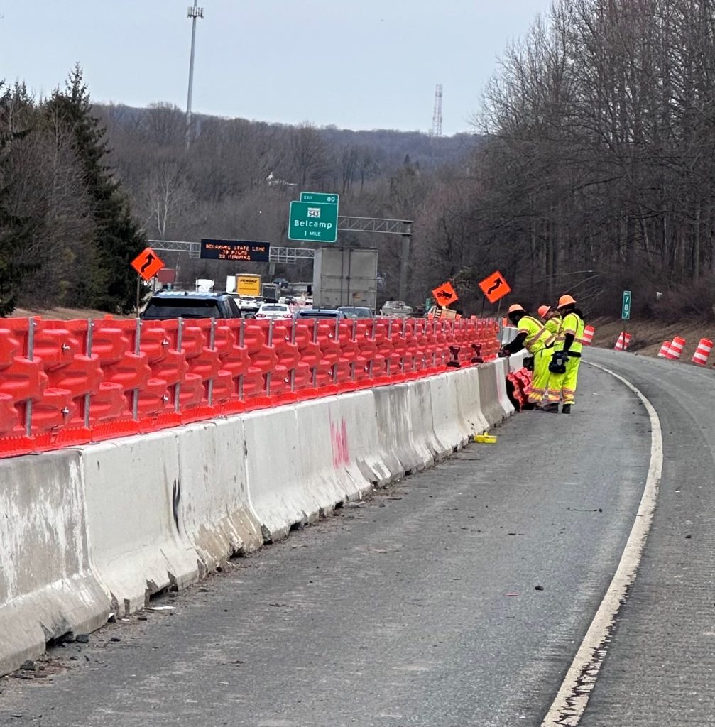 Road workers in neon gear work beside a highway lane closed by barriers and orange warning signs.