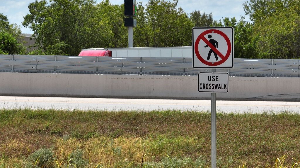 No pedestrian crossing sign above a “Use Crosswalk” sign near a road with grass and trees in the background.