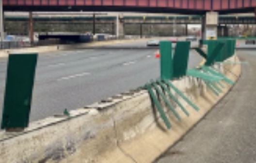 Damaged green metal fence along a highway barrier, some sections bent or missing, with empty lanes behind.